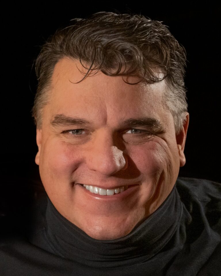 Eric Berlin smiling, in a black turtleneck against a dark background