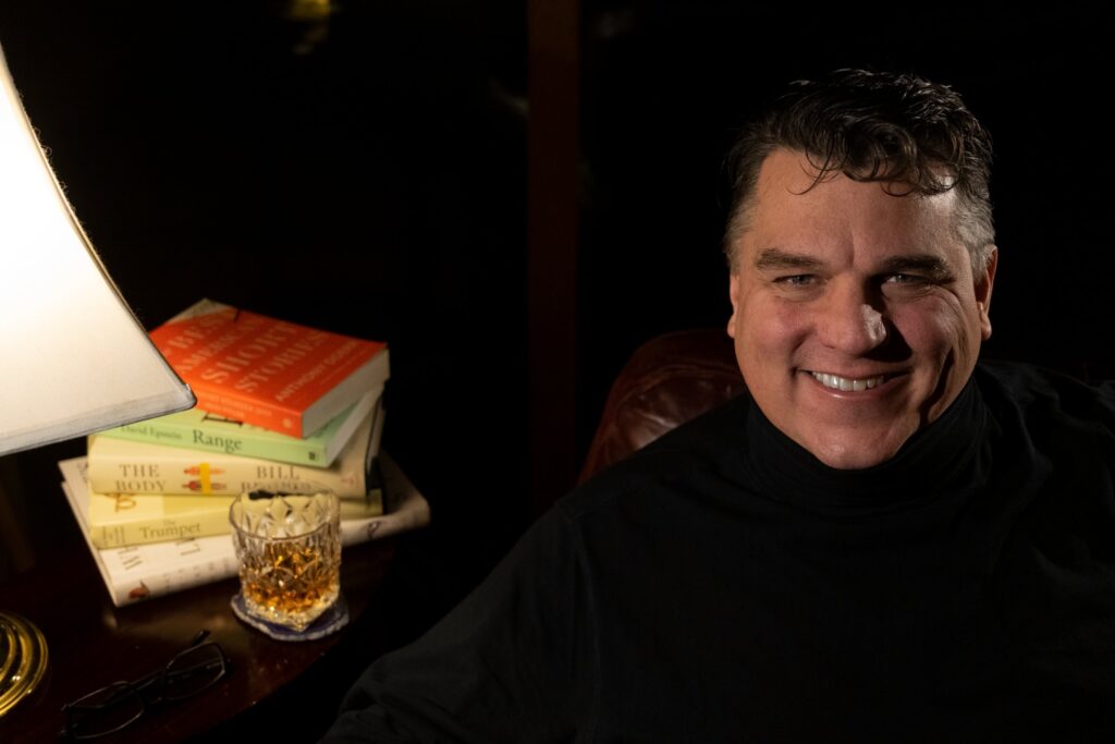 Eric Berlin smiling in a leather chair beside a lamp, a stack of books, reading glasses and a glass of whiskey